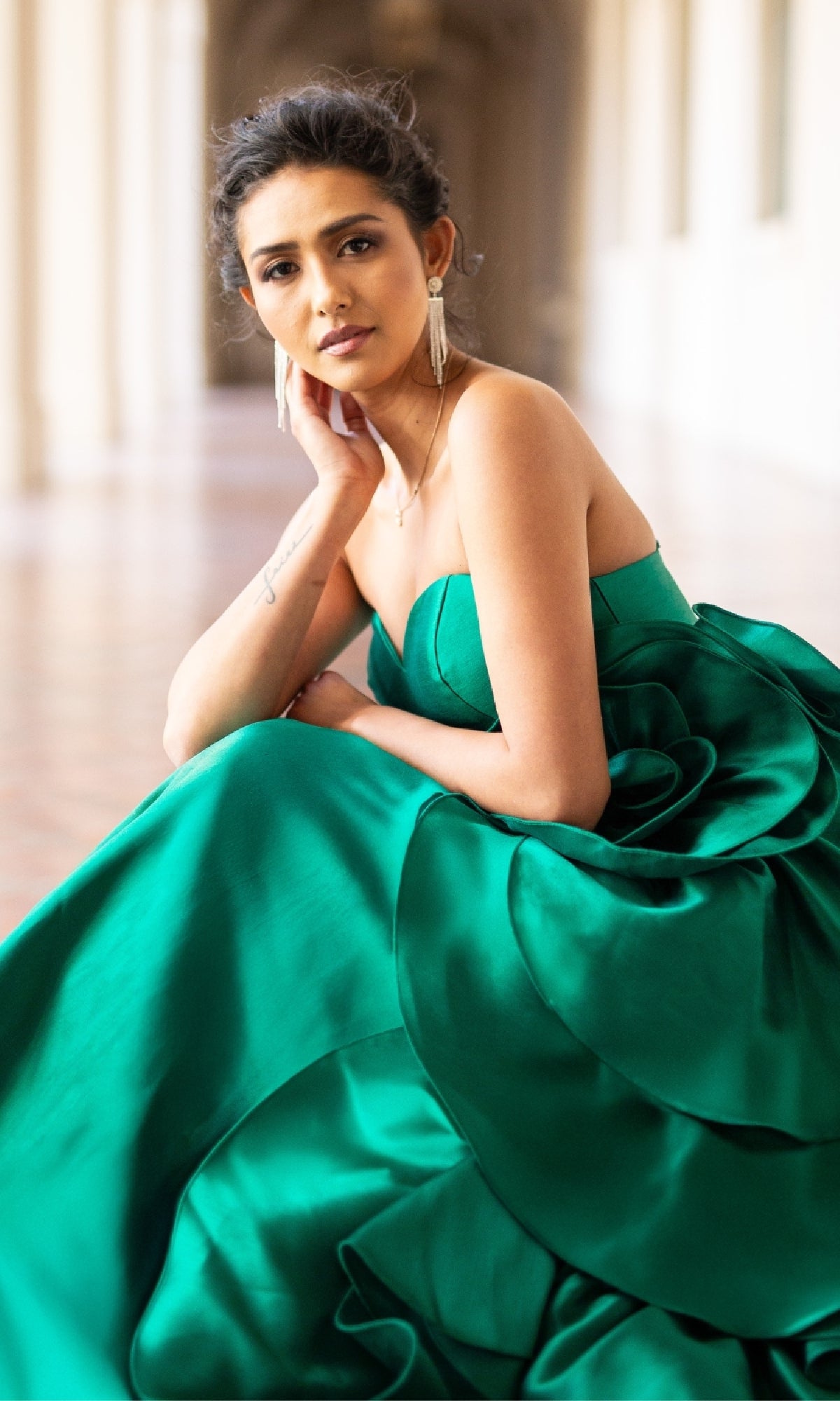 A woman in a Strapless Prom Ball Gown: Amelia Couture SU085, colored emerald green, sits on the hallway floor with her face resting on her hand, gazing at the camera.